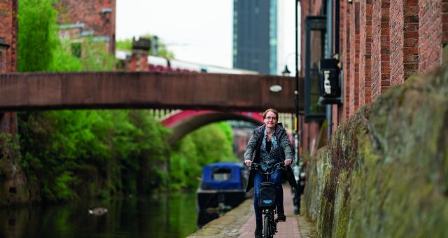 A woman cycles down a towpath along a canal in an urban setting