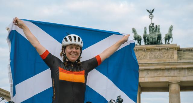 A woman stands holding a Scottish flag behind her while posing in cycling gear in front of the Brandenburg gates in Berlin, Germany