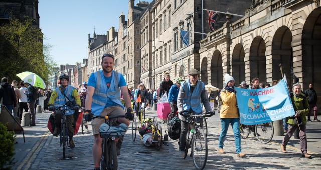 Many people are cycling down a cobbled street with grand buildings, including people on standard two-wheeled cycles and a three-wheeled handcycle. Two people are carrying a banner with a bicycle on it that reads 'This machine fights climate change'.