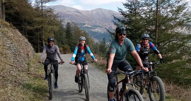 Four mountain bikers ride towards us on a gravel path in a mountainous landscape