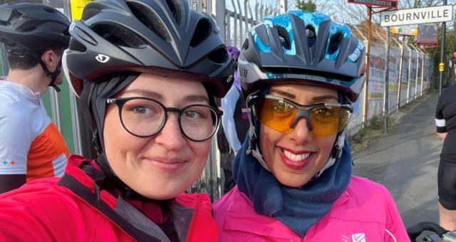 Two ladies pose for a selfie. They are wearing brightly coloured cycling jackets, glasses and helmets.