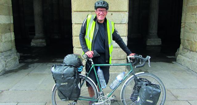 A man poses holding his bicycle which is loaded down with pannier racks front and back. He's wearing a high-vis jacket and a helmet