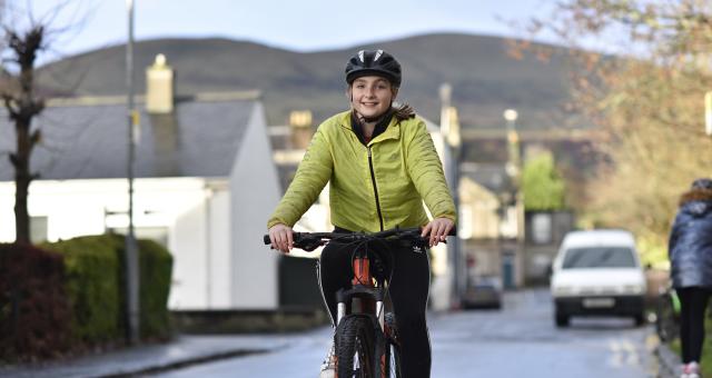 A girl wearing a yellow jacket and helmet is smiling as she cycles up the road. There are houses and hill in the background