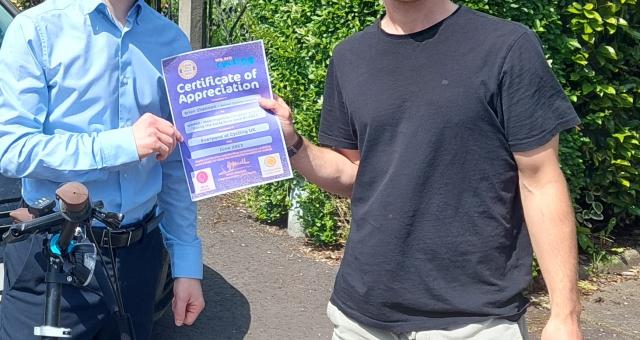 Two young men stand side by side in front of a garden hedge on a pavement. They are both holding a certificate