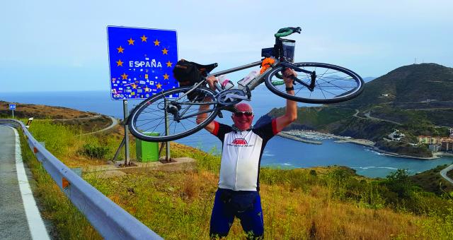 A older man wearing cycling clothes holds a road bicycle above his head in triumph along a coastal road. Behind him, a road sign with the EU flag on it surrounds the word 'Espana'.