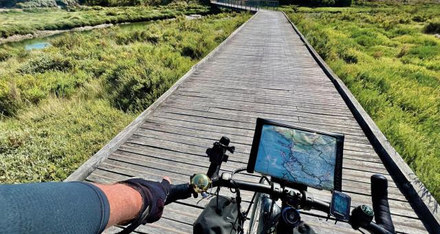 POV of a cyclist riding along a raised boardwalk above marshland. Their bike has a map attached to the handlebars and pannier bags on the forks