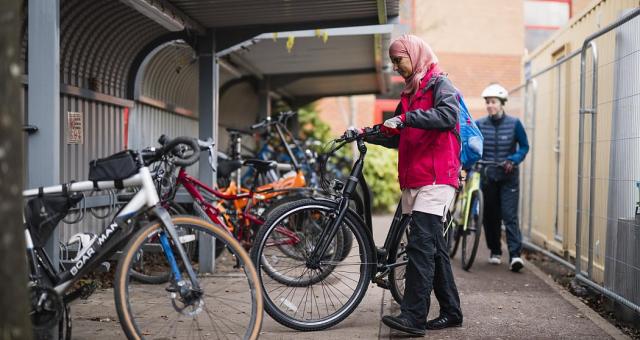 Woman wearing pink jacket and black trousers pushes e-bike into bike shed