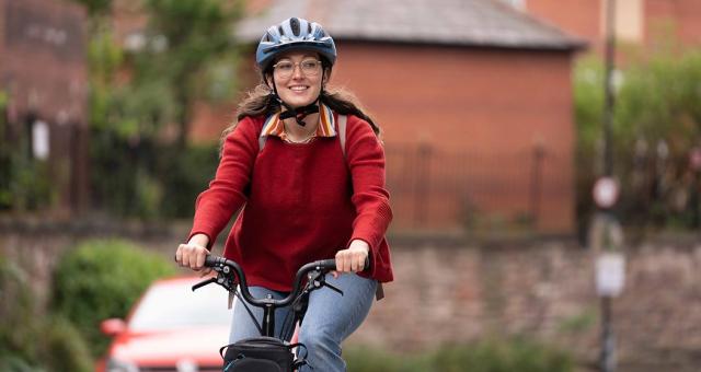A woman cycles along a road. She is wearing a red knitted jumper, a pair of jeans, a helmet and a pair of glasses. She is smiling