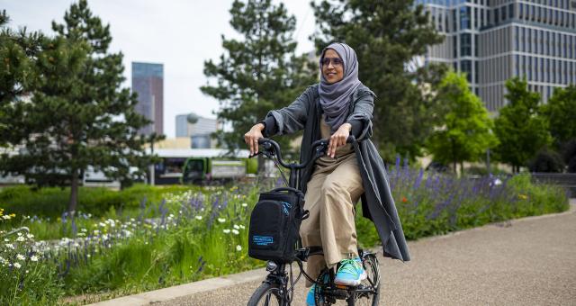 Woman pedals electric Brompton along cycle path with purple, white and yellow flowers and trees in background