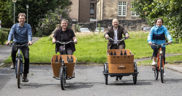 Active Travel Minister Patrick Harvie is riding a cargo bike, beside Suzanne Forup of Cycling UK who is riding a standard bike and representative from Bike for Good in Glasgow. The is a green space behind them