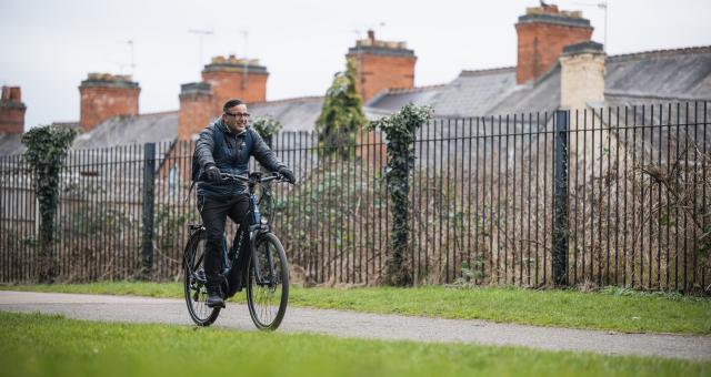 A man wearing glasses pedals a navy e-cycle through park in Leicester on a cloudy day