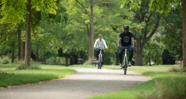 Two men pedal e-bikes through a park on a sunny day