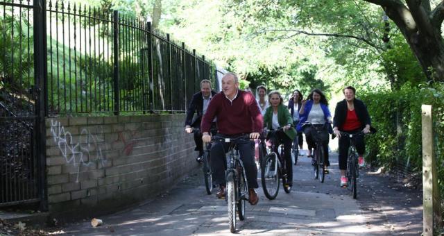 A group of people in normal clothes led by a man cycle down a path. There is a fence on the left with some greenery behind it