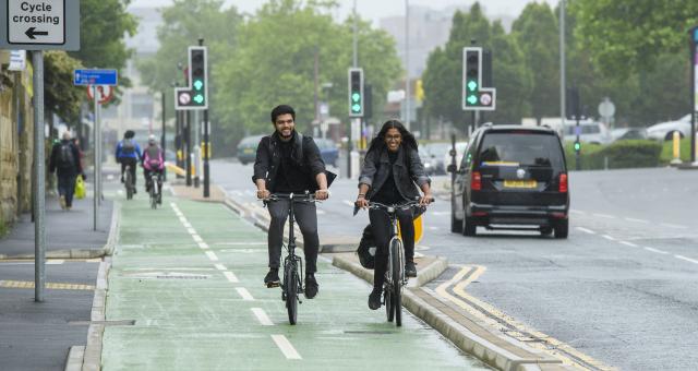 Two people ride side by side on a green cycle lane on a grey day in the city. Traffic lights on green in the background.