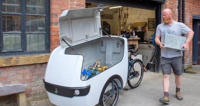 A stonemason wearing a grey t-shirt loads tools into a white e-cargo bike outside his workshop