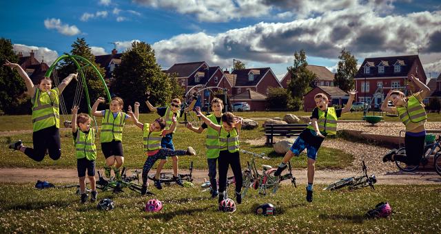 A group of children in hi-viz vests are jumping up in the air in a park next to their bikes on the ground