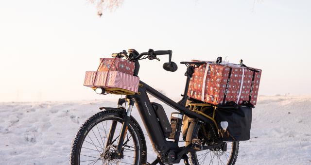 A black e-bike is loaded up with wrapped presents on the front and rear racks. It's propped up in a snowy landscape.