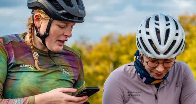 Two women wearing cycling jerseys and helmets. One is looking down, the other has a phone in her hand