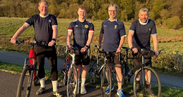 Four men are standing with their bikes on a paved path. They’re wearing Scotland football shirts and black shorts, one is wearing black tracksuit bottoms. Two bikes are hybrids, two are road bikes.