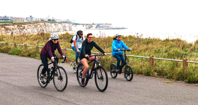 Four people are cycling along a paved path on a clifftop with a seaside town in the background. They're on a mix of bikes, including road, hybrid and folding. They're wearing normal clothes.