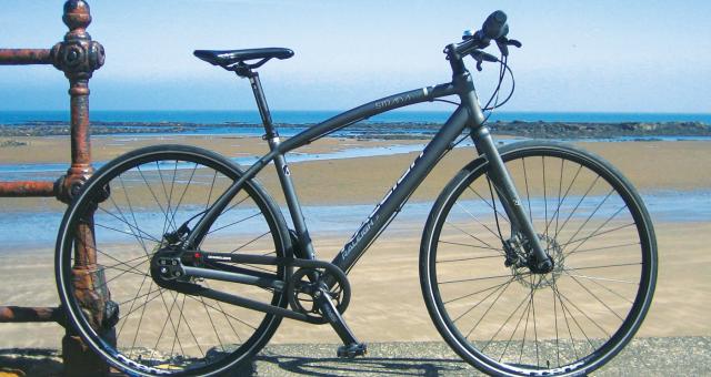A dark grey hybrid bike with flat handlebar, disc brakes and no mudguards. In the background is a beach with the tide far out