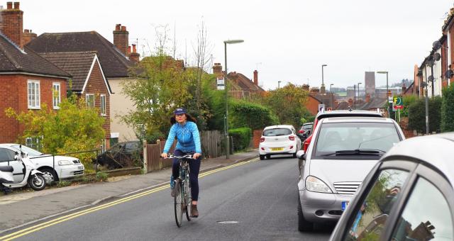 A woman is riding along a road in the centre of the lane so she can be seen. She is wearing a cap, blue jersey and blue leggings