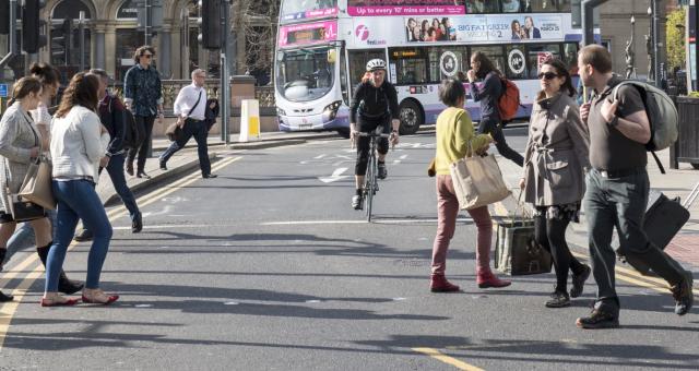 A woman is cycling on a busy city centre road. People are crossing in front of her and behind her is a bus