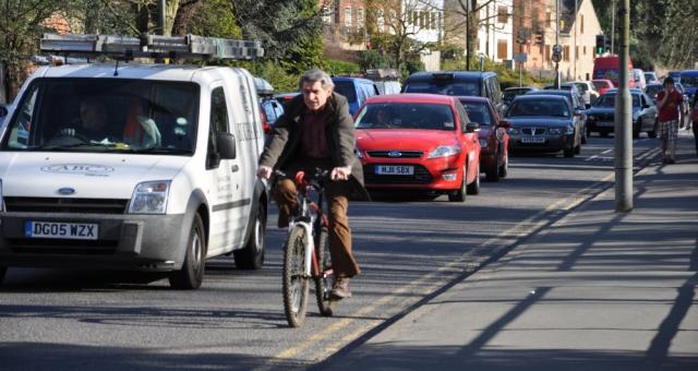 A man is cycling along a busy road with a line of traffic to his right