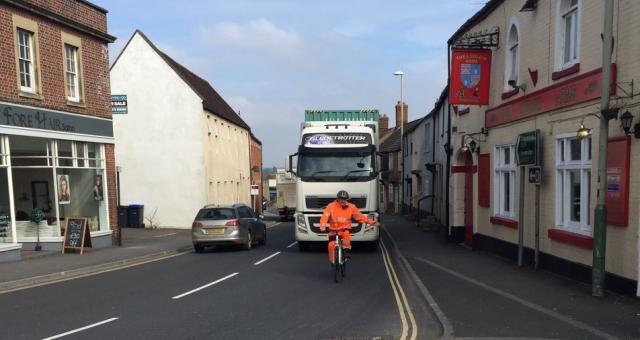 A man in an orange hi-vis outfit is cycling in front of a lorry. He is indicating to go left