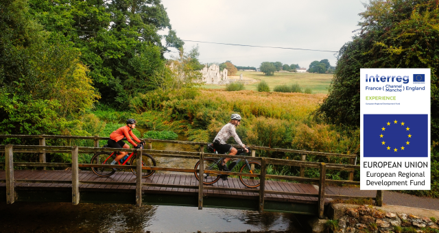 Two people cycle across a wooden bridge with a ruined abbey peeking through the trees behind