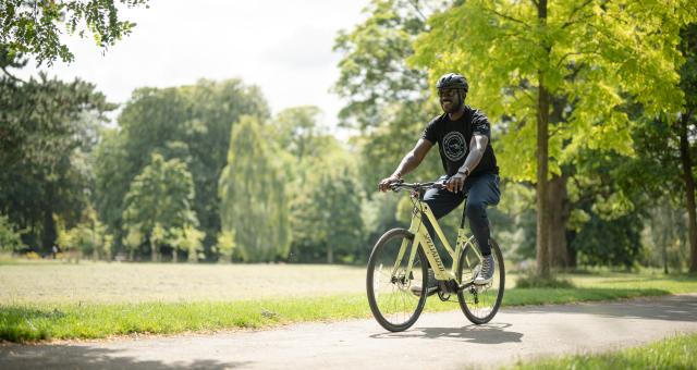 A wearing a black t-shirt and helmet pedals an e-cycle through a park on a sunny day