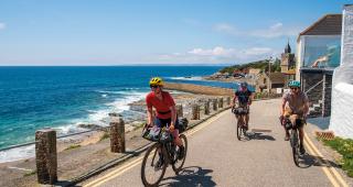 Three people are cycling along a single-lane tarmac road next to the sea