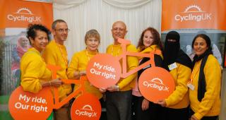 A group of people all wearing knitted yellow sweaters are standing together in front of Cycling UK branded boards and holding cardboard bikes with the words 'My Ride. Our Right' in one wheel and the Cycling UK logo in the other
