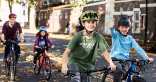 Two young boys are riding their bikes with a woman and a young girl cycling behind them, all on a segregated cycle lane. One of the boys is giving the camera a thumbs up