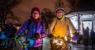 Two women stand smiling at the camera on a dark street with bikes lit up with fairy lights