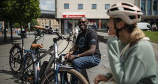  A man in jeans and T-shirt and a woman in a sweater and cycling helmet are sitting on a wall in an urban setting. Two bikes are leaning against the wall with them