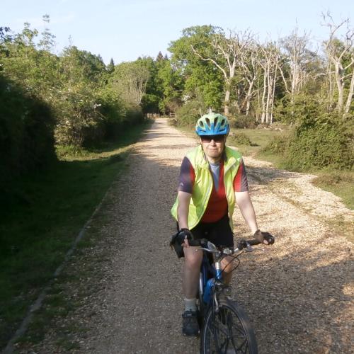 A woman is cycling towards the camera. She is wearing black shorts, an orange and grey jersey and yellow hi-vis jacket, along with a blue and green helmet and sunglasses. The bike is blue. She is cycling on a gravel path bordered by woodland on both sides