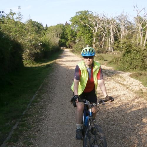 A woman is cycling towards the camera. She is wearing black shorts, an orange and grey jersey and yellow hi-vis jacket, along with a blue and green helmet and sunglasses. The bike is blue. She is cycling on a gravel path bordered by woodland on both sides