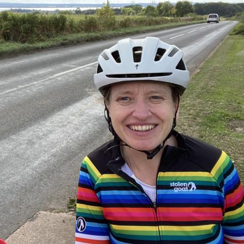 A woman is standing on a grass verge at the side of a road. She is wearing a white cycle helmet and a bright, stripey Stolen Goat cycling jersey. She is smiling at the camera.
