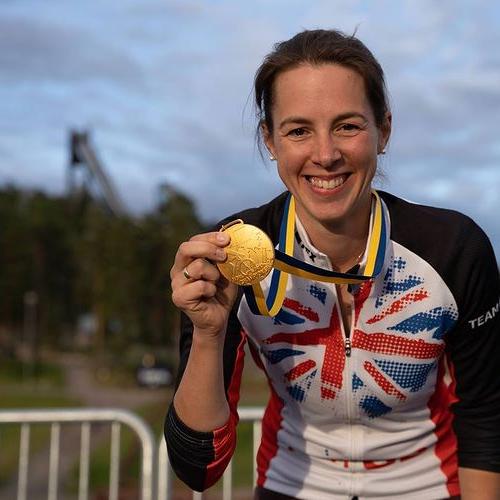 A woman in a GB cycling jersey is holding up a gold medal and smiling to the camera