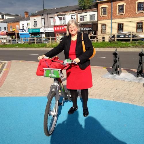 A woman is standing with a hire bike on some of Birmingham’s cycle infrastructure, the blue routes. She is wearing a red dress, black cardigan and black tights. She has a yellow handbag over her shoulder and a red bag in the bike’s basket