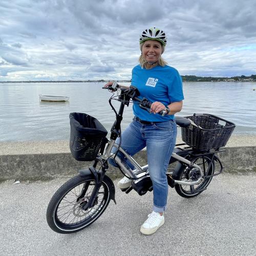 Lucie on a silver e-cycle with front and back baskets, standing in front of Poole harbour. She is wearing blue jeans and a blue BH Active Travel T-shirt