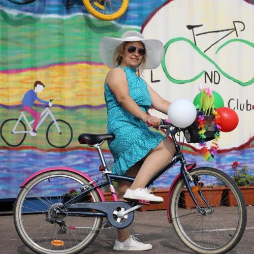 A smiling woman with long blonde hair and sunglasses is standing astride a black and pink bike. She is wearing a blue dress and white sun hat. The bike’s basket is decorated with balloons and plastic flowers