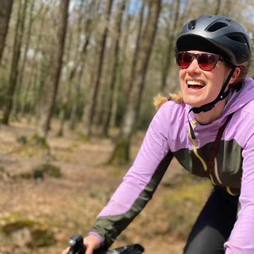A woman in a lilac and forest green cycling jacket and silver helmet is cycling off road through a forest. It’s autumn and the trees are bare, there are leaves all over the forest floor