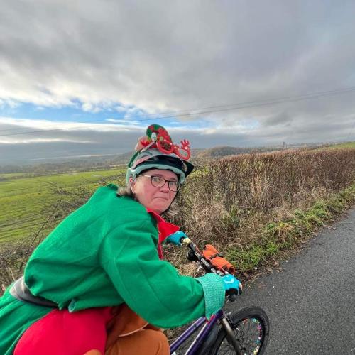 A woman is riding a bike while wearing a Christmas elf outfit. She is looking back at the camera and pulling a funny face