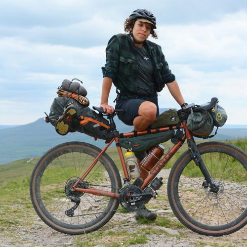 A woman is posing with a heavily laden bikepacking bike. She is wearing black shorts, a black Rapha T-shirt and check shirt, and a black cycling helmet with a cap underneath. She is in a remote location with mountains in the background