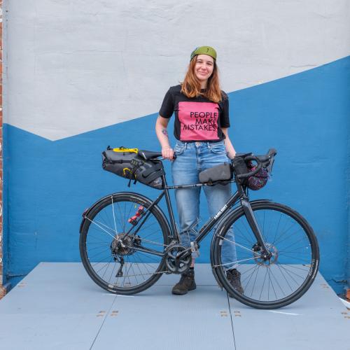 Adna stands smiling with her black bike. She is wearing a cap, black and pink slogan t-shirt and jeans. 