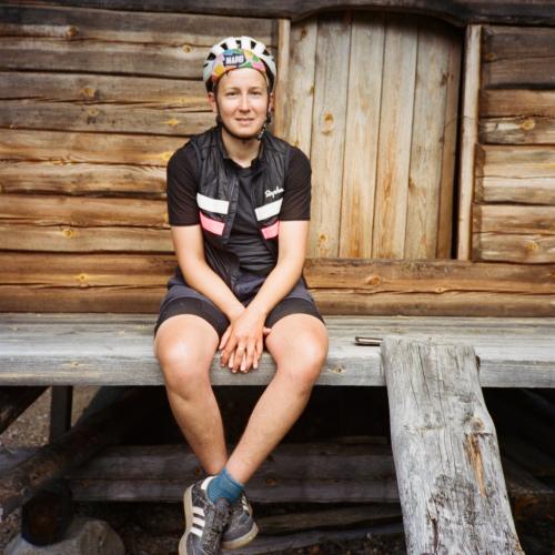 Anna sits on a wooden veranda. She wears a helmet, black top, gilet, shorts and trainers.