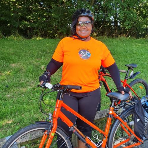 Joscelyne smiling with her red bike. She is wearing an orange Sara Park CCC t-shirt, black leggings and helmet.