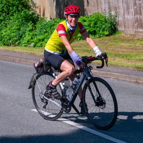 Yasmin smiling as she rides a white Trek bike on road. She is wearing a red helmet, yellow gilet over a red jersey and black cycling shorts. 